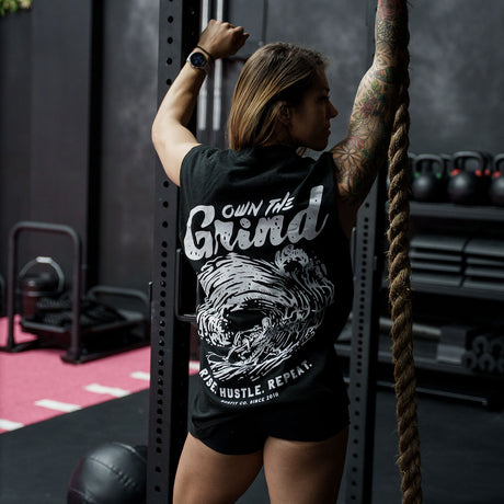 Female model in a gym wearing sleeveless Street Tee with 'Own The Grind' graphic